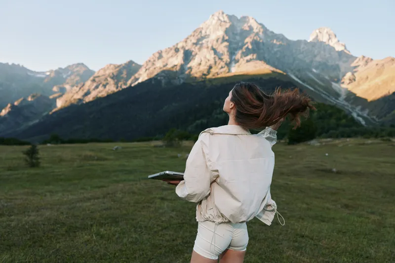 A woman stands in a wide open field with distant mountains, gazing toward the horizon, wearing a light jacket and shorts, capturing a moment of outdoor freedom and adventure.