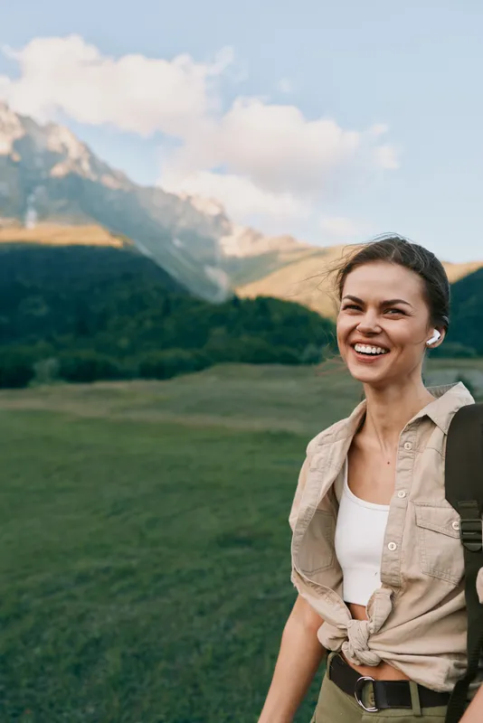 Young woman with dark hair stands in a sunlit mountain meadow smiling and enjoying a carefree outdoor moment in nature and fresh air.