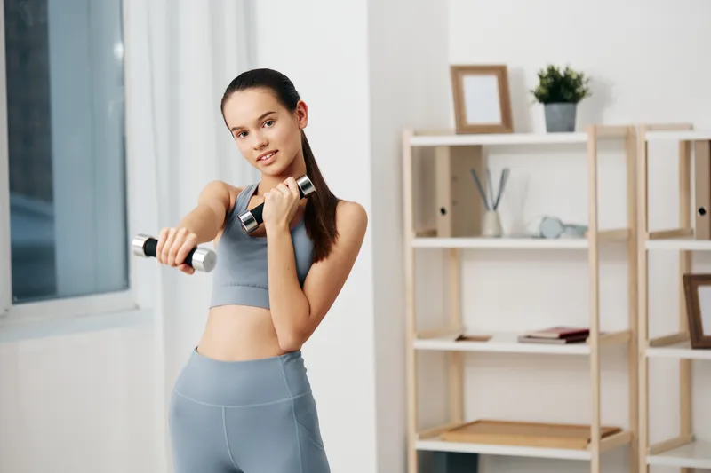 Female fitness enthusiast in athletic wear lifts dumbbells in a bright home gym, focused expression, minimalist shelves and clean space behind.