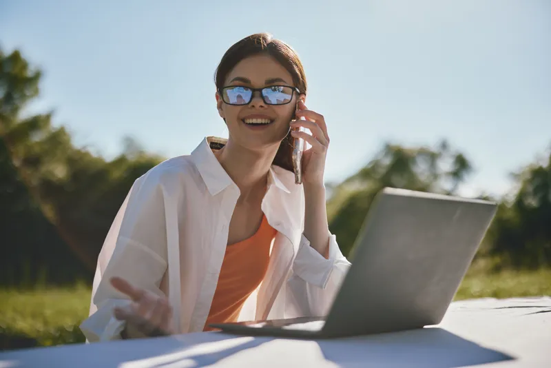 Young woman enjoying a sunny day while working on her laptop, wearing sunglasses and a casual outfit outdoors