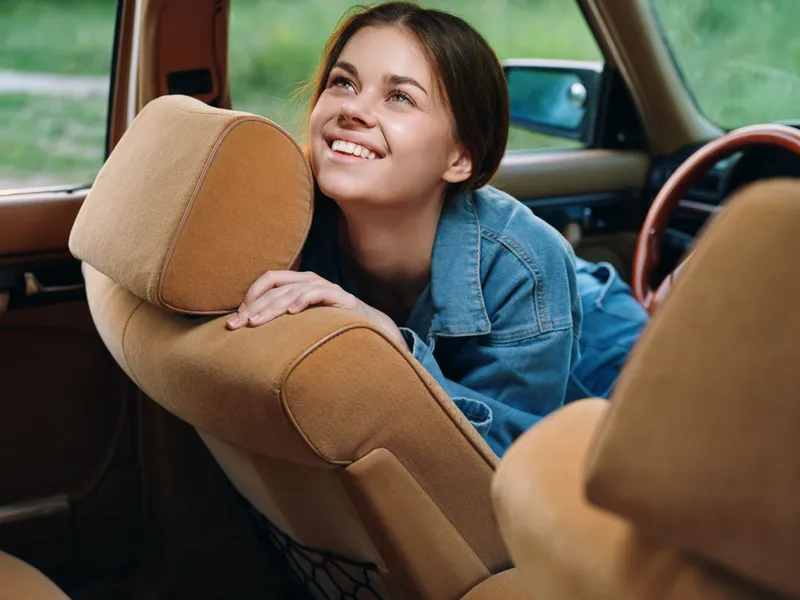 Young woman with a joyful expression in a vintage car, wearing a denim jacket, enjoying a sunny day in nature with vivid green background.