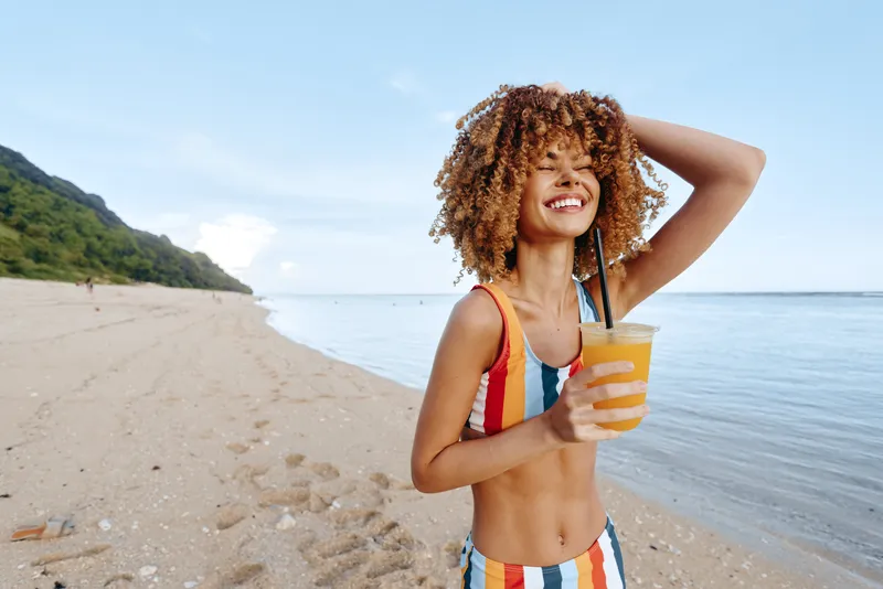 Bright beach scene with a smiling woman in a striped swimsuit holding a cold orange drink, sunlit sand and calm ocean, lush coastline behind, a carefree summer mood