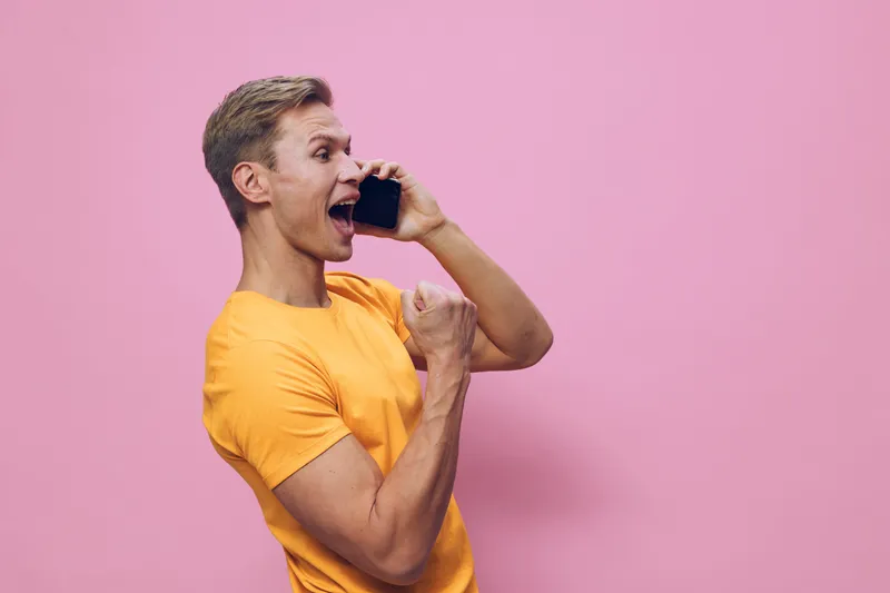 Cheerful young woman with short blond hair wearing a bright yellow t-shirt talking on smartphone smiling thrilled excited in studio with pink background. People lifestyle concept