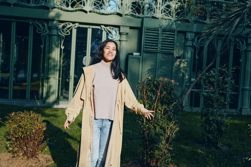 Woman wearing a long beige coat over a light sweater and denim in a sunny garden, standing near an ornamental gate, casual outdoor fashion with a relaxed, confident pose