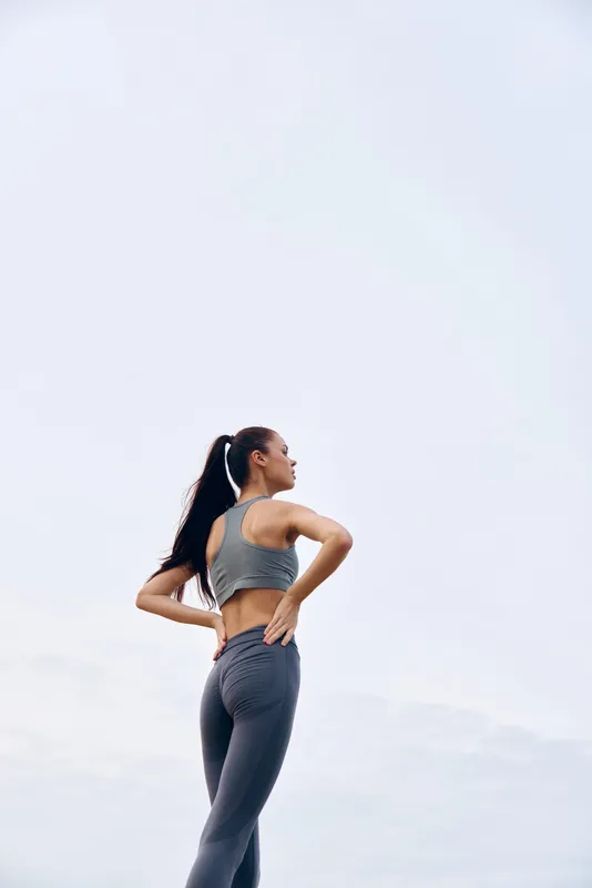 Athletic woman in gray activewear enjoying workout outdoors, demonstrating strong posture and determination in a bright sky environment during warm weather