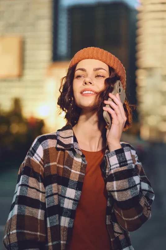 woman phone urban portrait smile beanie candid lifestyle: Young woman on phone in city at golden hour, warm golden hour glow, authenticity, mindful living and emotional storytelling in street scene