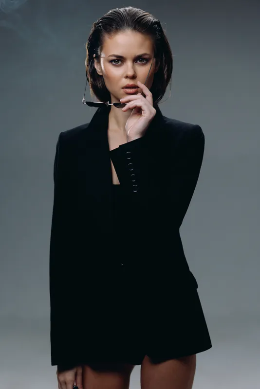 Stylish woman in black blazer posing confidently with wet hair and intense look against gray background