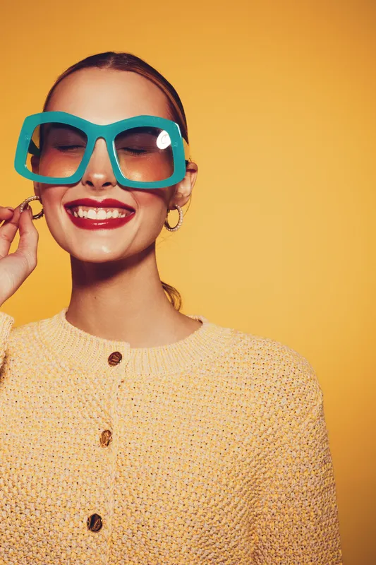 Smiling woman with large turquoise sunglasses and red lipstick poses against yellow background. She wears a beige knitted sweater and gold hoop earrings. Concept of fashion and joy.