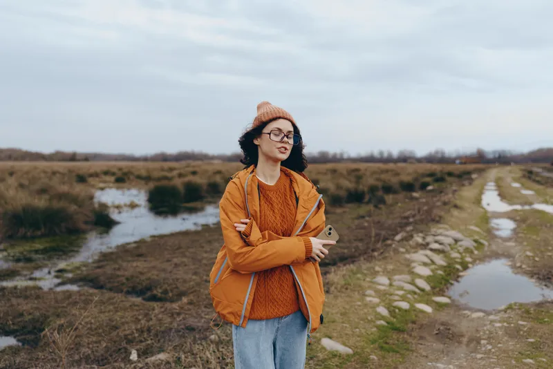 Woman lifestyle outdoors countryside jacket, arms crossed, stream path, cloudy sky, autumn walk, confident smile, orange jacket and glasses, rural landscape, soft breeze, cool daylight