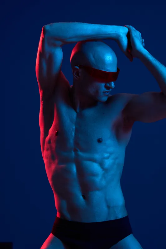 Athletic man posing in dynamic stance, showcasing toned physique and expressive gestures under vibrant blue and red lighting for a striking visual impact.