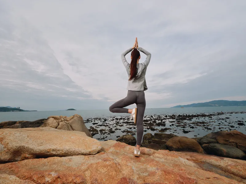 Young woman practicing yoga on a rocky beach, showing tranquility and focus, wearing a gray top and leggings, with a serene ocean background at sunrise