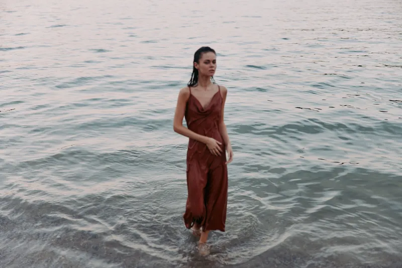 Woman stands in shallow sea water wearing a rust red dress, barefoot as gentle waves lap her ankles, while soft light highlights the scene with a calm coastal mood.