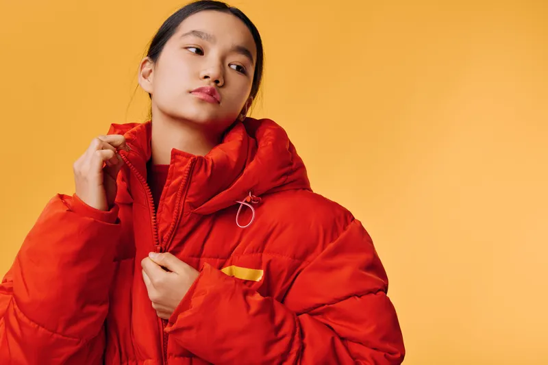 Young woman wearing bright red puffer jacket posing against yellow background with confident expression. Studio portrait of casual winter fashion style with warm outerwear for cold season.