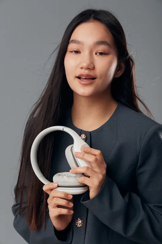 Woman holding wireless headphones with hands in studio, long hair and dark jacket on grey background, technology device for audio listening and casual style closeup portrait