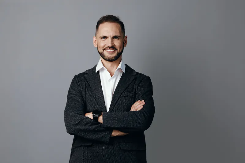 A professional man stands on a solid colored backdrop with arms folded, projecting confidence and warmth. His poised smile communicates trust, ambition, and readiness for success.