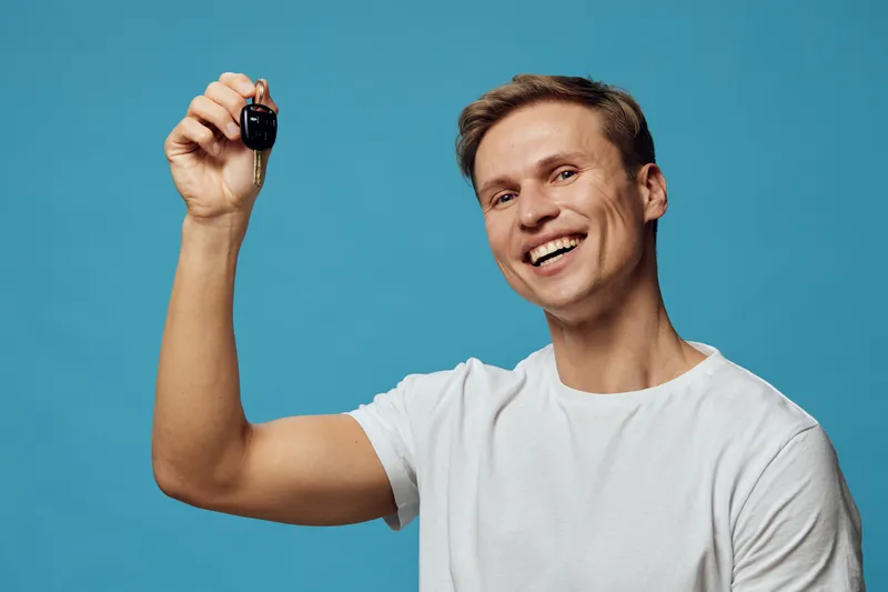 Smiling young man with short brown hair wearing white t-shirt holding car key in hand on blue background studio portrait. People lifestyle concept, success, achievement, joy, automobile, motivation.