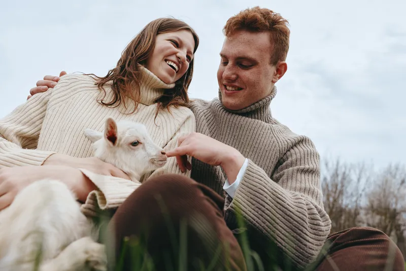 A couple outdoors share a joyful moment with a friendly goat kid on a sunny day, relaxed in sweaters, smiling, sitting on grass, close and affectionate, creating a warm rustic lifestyle scene and