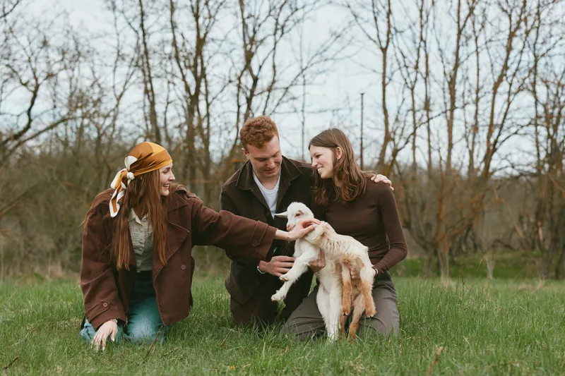 Three friends outdoors with a playful goat kid in a grassy field, sharing smiles. Casual outfits, soft natural light, rural vibe. A candid lifestyle moment highlighting companionship, nature, and