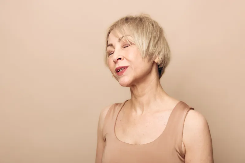 Portrait of a mature woman with short blonde hair smiling gently with closed eyes on beige background. Concept of natural beauty, aging gracefully, and calm self-acceptance.