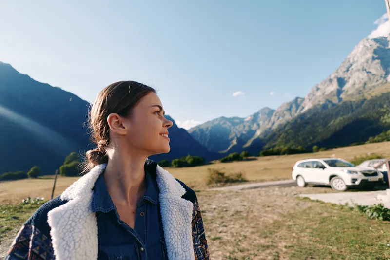 Woman portrait in alpine mountains near parked car, landscape and nature scene for travel and outdoors adventure, young traveler looking sideways with clear sky and valley backdrop