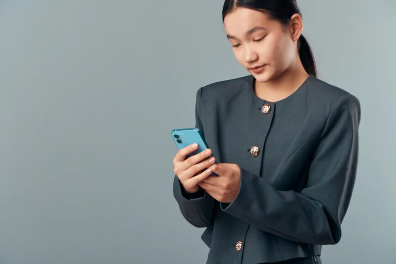 Woman wearing dark jacket using smartphone with focused expression on gray background. Portrait of young female texting or browsing online with mobile device in casual pose.