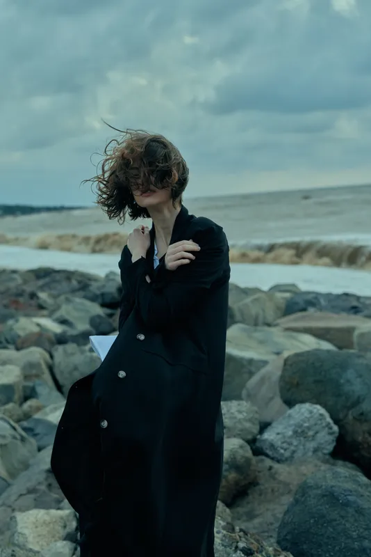 Coastal moody scene with a woman in a long dark coat on rocky shore, wind in hair, overcast sky, cinematic fashion mood, solitary figure by rough sea
