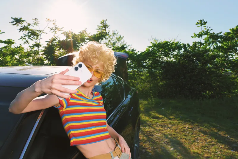 Cheerful woman outdoors leaning on a car, striped top, bright sun, casual playful selfie, retro film colors, carefree lifestyle, lush greenery background bright sunlit vibrant mood