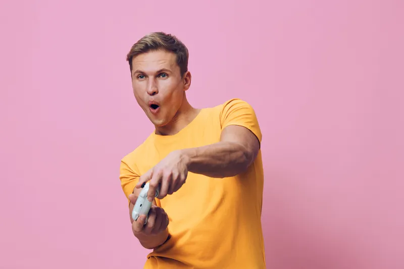 surprised man playing video game holding controller in yellow t-shirt isolated on pink background casual lifestyle concept
