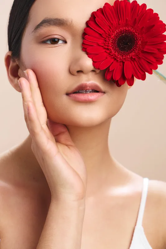Asian woman gazes softly at the camera, a bold red flower beside her cheek, creating a serene beauty portrait that highlights elegance, calm and natural charm.