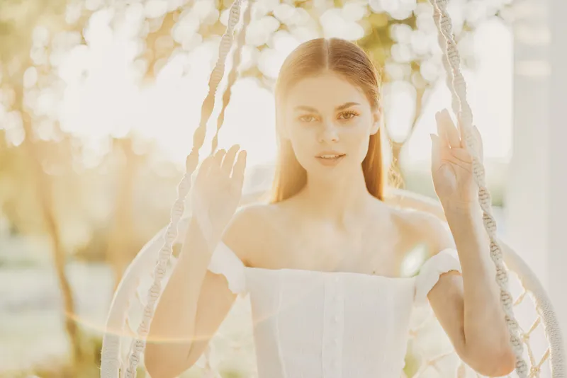 Young woman in a white dress on a swing, with a soft sunny background, conveying a serene and joyful mood in a beautiful outdoor setting