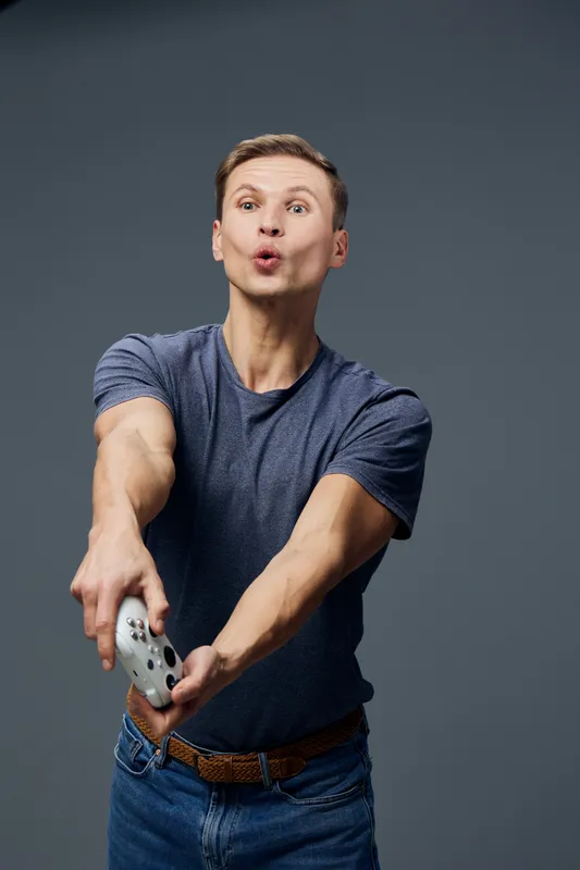 Man playing video game, holding a game controller, making a playful face with pursed lips, casual blue t-shirt, modern studio background, fun entertainment activity, indoor setting, people lifestyle c