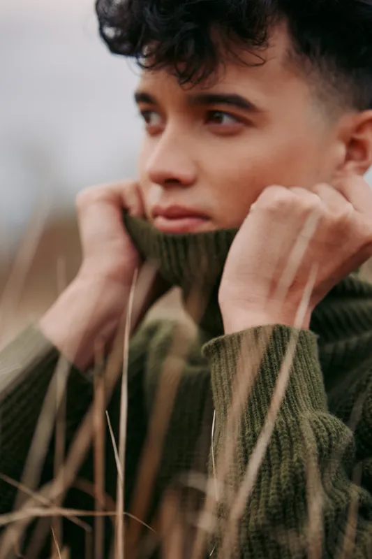 Close candid portrait of a young man pulling a knit sweater collar up against the wind, conveying natural authenticity and quiet reliability in a soft earthy outdoor moment.