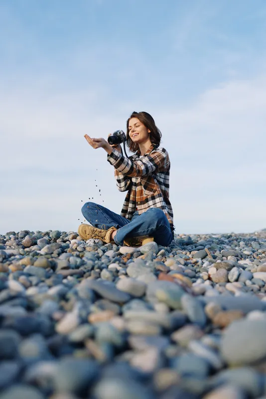 Woman sits on a pebble beach, outdoors and casual, playfully throwing small stones into the air, capturing a joyful moment under clear sky with calm sea and relaxed vibe.