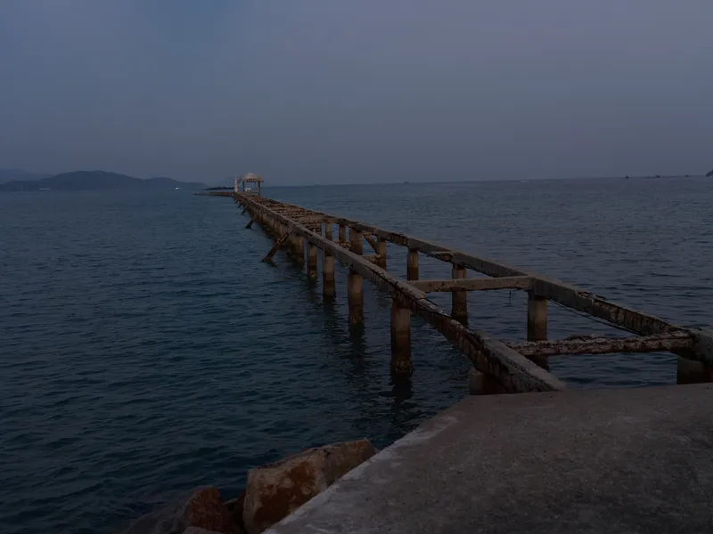 Rusty old pier extending into the calm sea during twilight, featuring deep blue tones and a serene coastal atmosphere.