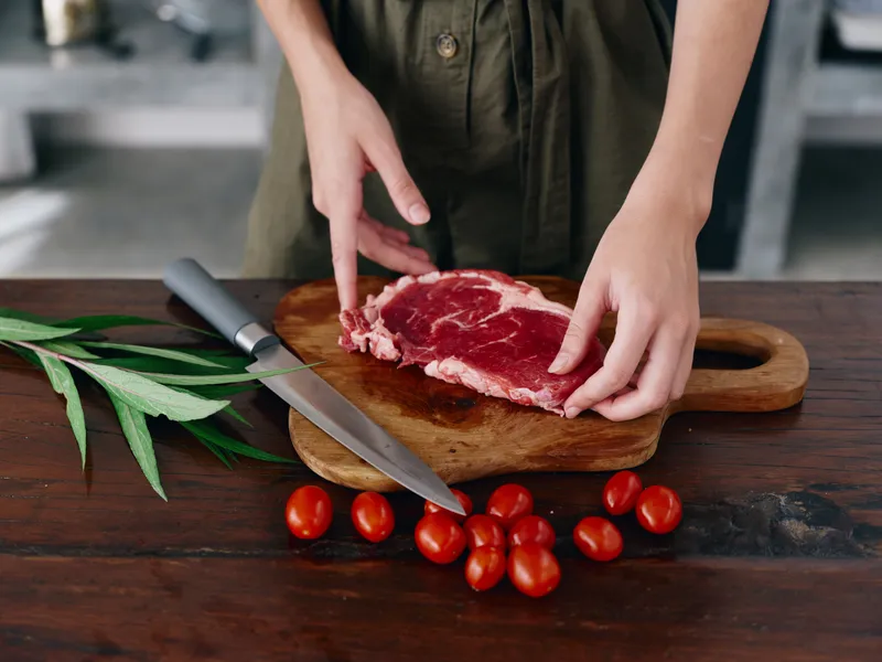 Женщина с ножом в руке нарезает свежее стейковое мясо для жарки на кухне