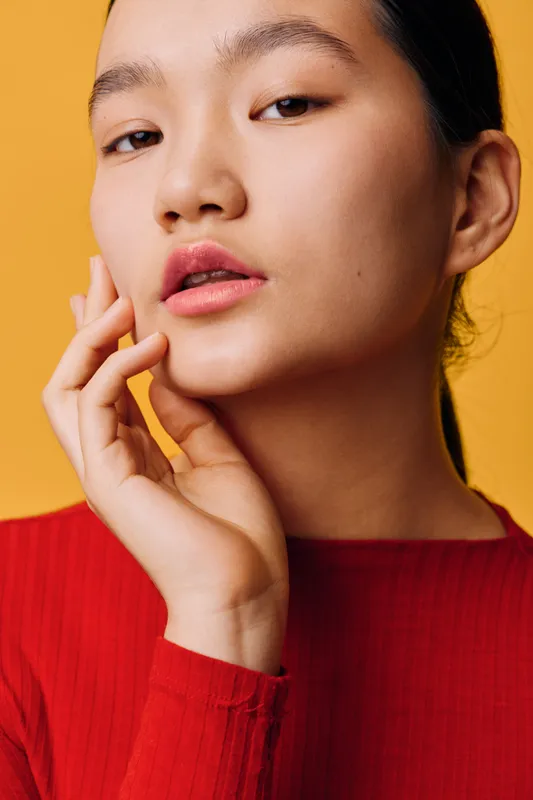 Young woman with natural makeup posing with fingers near face looking at camera on yellow background. Female model wearing red top with smooth skin and parted lips in studio lighting.