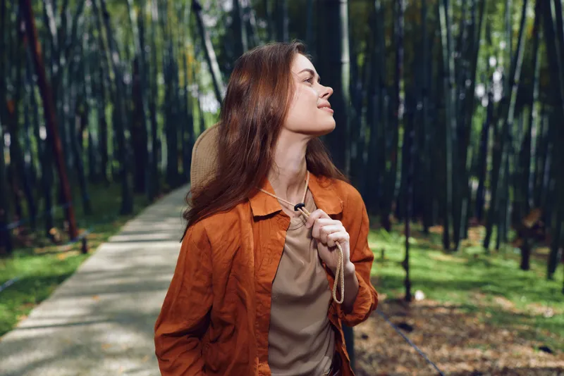 Woman in forest on a paved path, travel portrait of a happy outdoors explorer breathing fresh air, wearing orange jacket and backpack strap, enjoying nature walk.