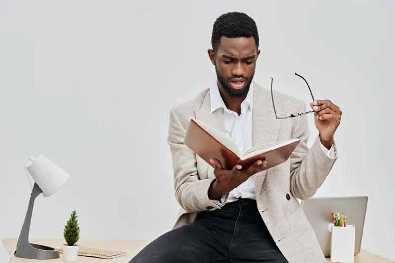 Thoughtful man reading a book and adjusting glasses in modern indoor setting, casual attire, neutral background, focused expression, lifestyle and leisure concept, studio shot.