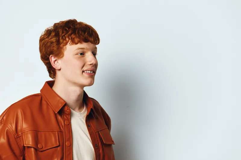 Young man with red hair in brown leather jacket looks sideways with slight smile on light background. Casual style, portrait of male model with curly hair for fashion or lifestyle.