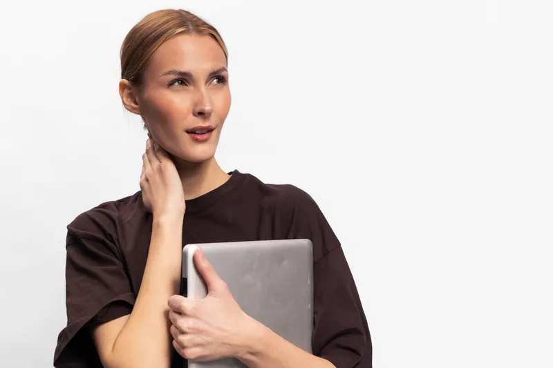 Thoughtful woman with brown hair in black shirt holds laptop and touches neck, looking away on white background. Concept for technology, contemplation, and casual work lifestyle.