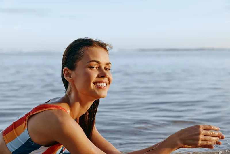 Smiling woman in striped life jacket sits by the ocean shoreline at sunset, relaxing with sunscreen lotion, calm waves behind, summer seaside lifestyle concept for wellbeing and travel