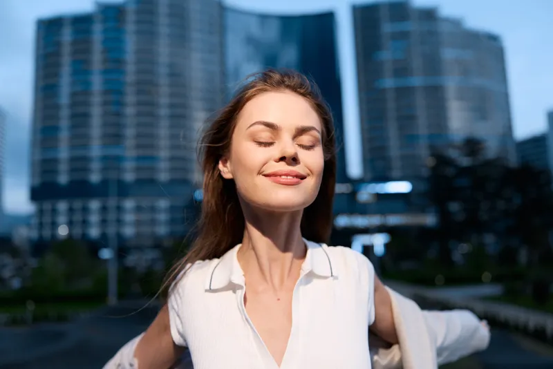 Businesswoman enjoying a moment of serenity in a modern urban environment, radiating confidence and positivity with a bright smile, dressed elegantly in a white blouse.