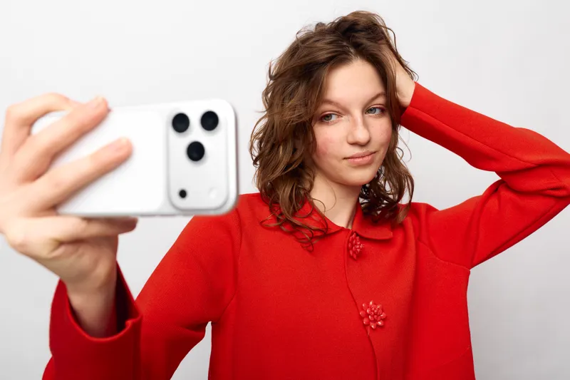 Young woman in red sweater takes a selfie with smartphone, posing with hand in hair on white background. Concept of casual style, social media, and self-portrait photography.