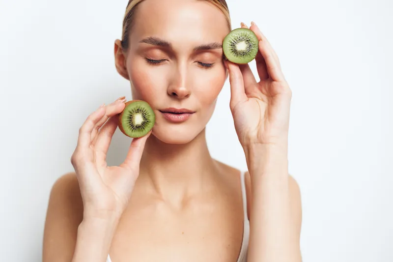 A young woman with clear skin holds two halves of a kiwi fruit near her face with closed eyes. The bright studio background highlights natural beauty and healthy lifestyle concepts.