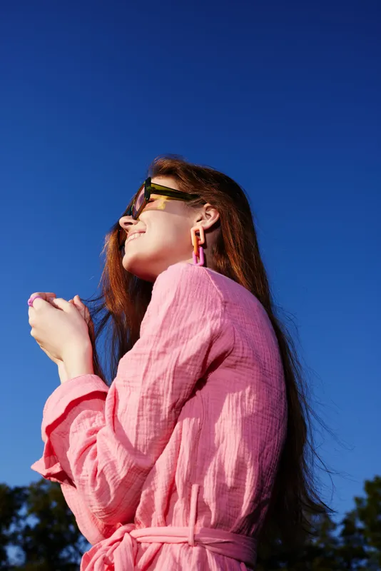 Young woman in pink dress enjoying a sunny day outdoors, smiling with coffee in hand, showcasing happiness and relaxation under a clear blue sky