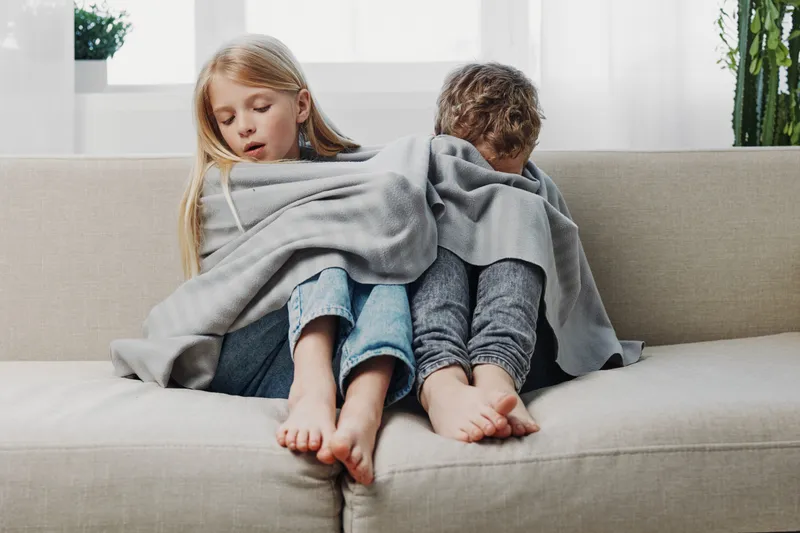 Two children wrapped in a grey blanket sit back to back on a beige sofa in a bright room. The scene conveys comfort, warmth, and sibling connection in a cozy home environment.