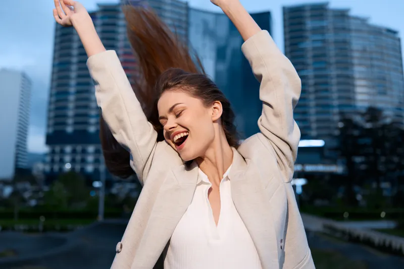Business woman expressing joy in an urban setting, dressed in a light blazer, with hair flowing in the wind against a modern cityscape backdrop.