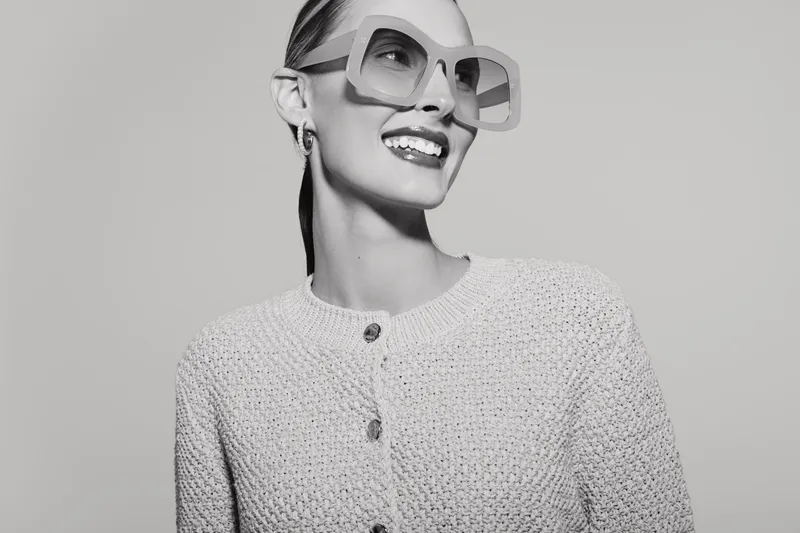 Smiling white woman with fair skin in large sunglasses and textured sweater looks up. Studio portrait in black and white with positive mood and fashion concept.