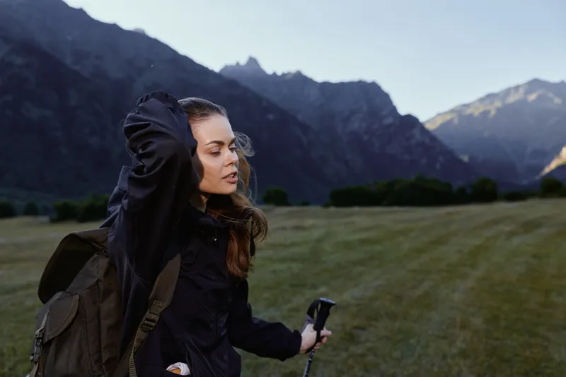 A young woman hiker stands in a vast meadow beneath towering mountains, adjusting her hair with a thoughtful gaze, ready for an outdoor trek and fresh alpine air.