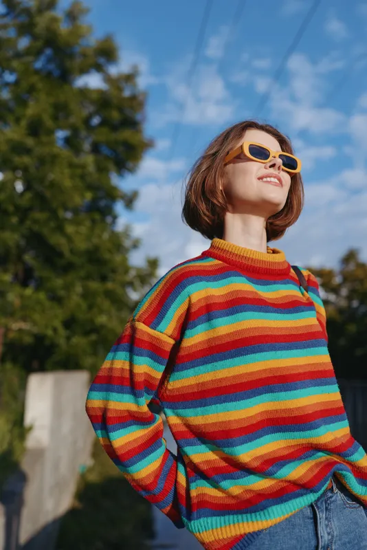 Young woman in a striped sweater and yellow sunglasses smiling outdoors, casual outfit and bob haircut, looking up under sunny sky in urban park for lifestyle travel portrait.
